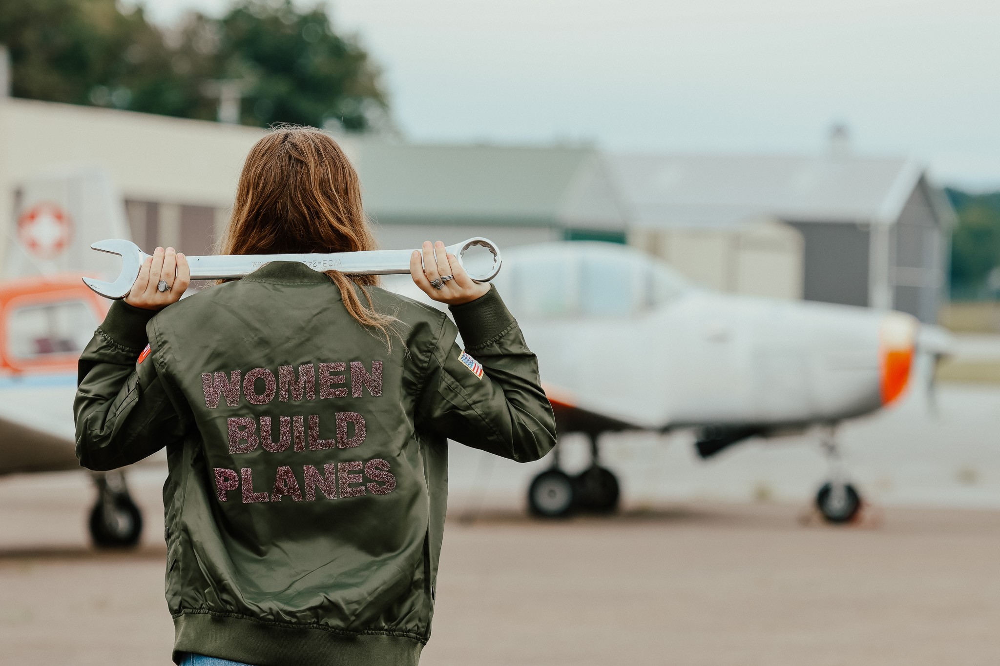 A woman in a flight jacket standing in front of a plane with a wrench over her shoulder.
