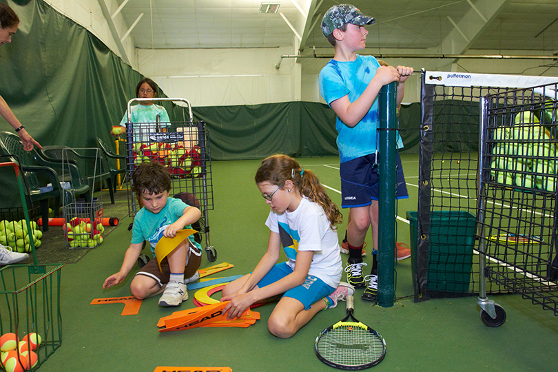A group of kids sit on a tennis court with racquets and tennis balls.