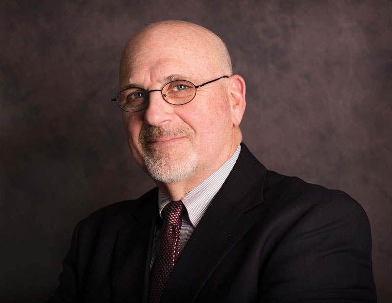 a man in a dark suit and tie with a bald head, facial hair, and glasses