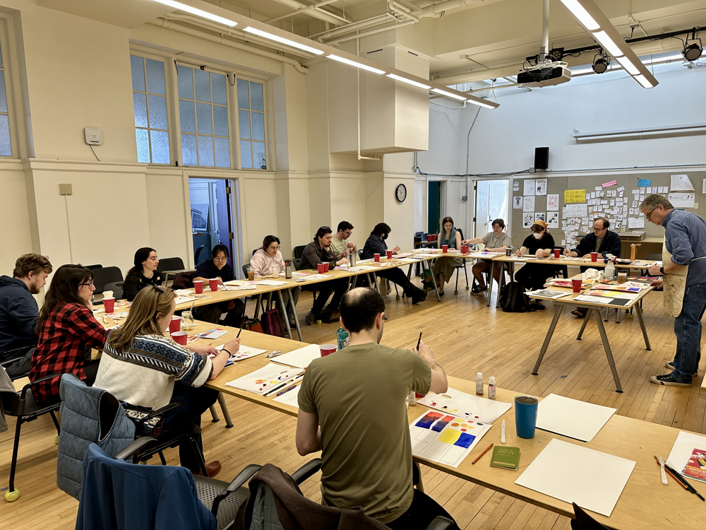 a group of people seated at tables set up in a u shape and painting