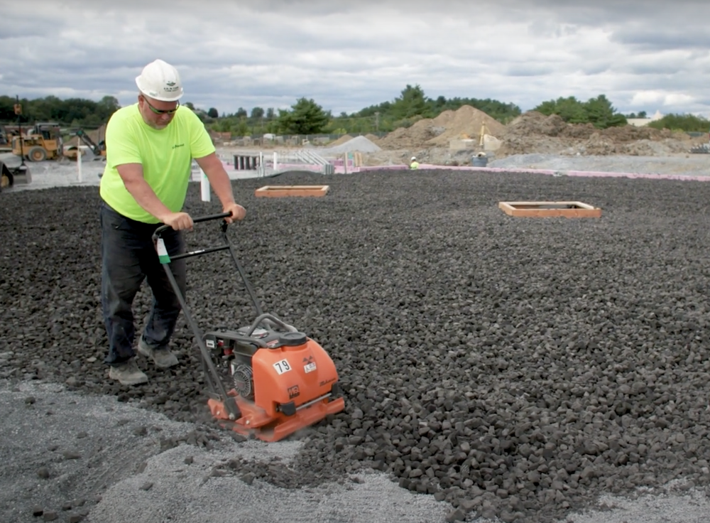a man in a bright yellow shirt wearing a hard hat and working with construction equipment to tamp down materials at a construction site foundation
