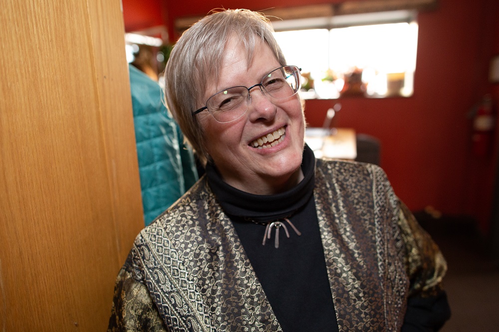 a woman with short hair, glasses, and a big smile, wearing a black & gold jacket and black shirt