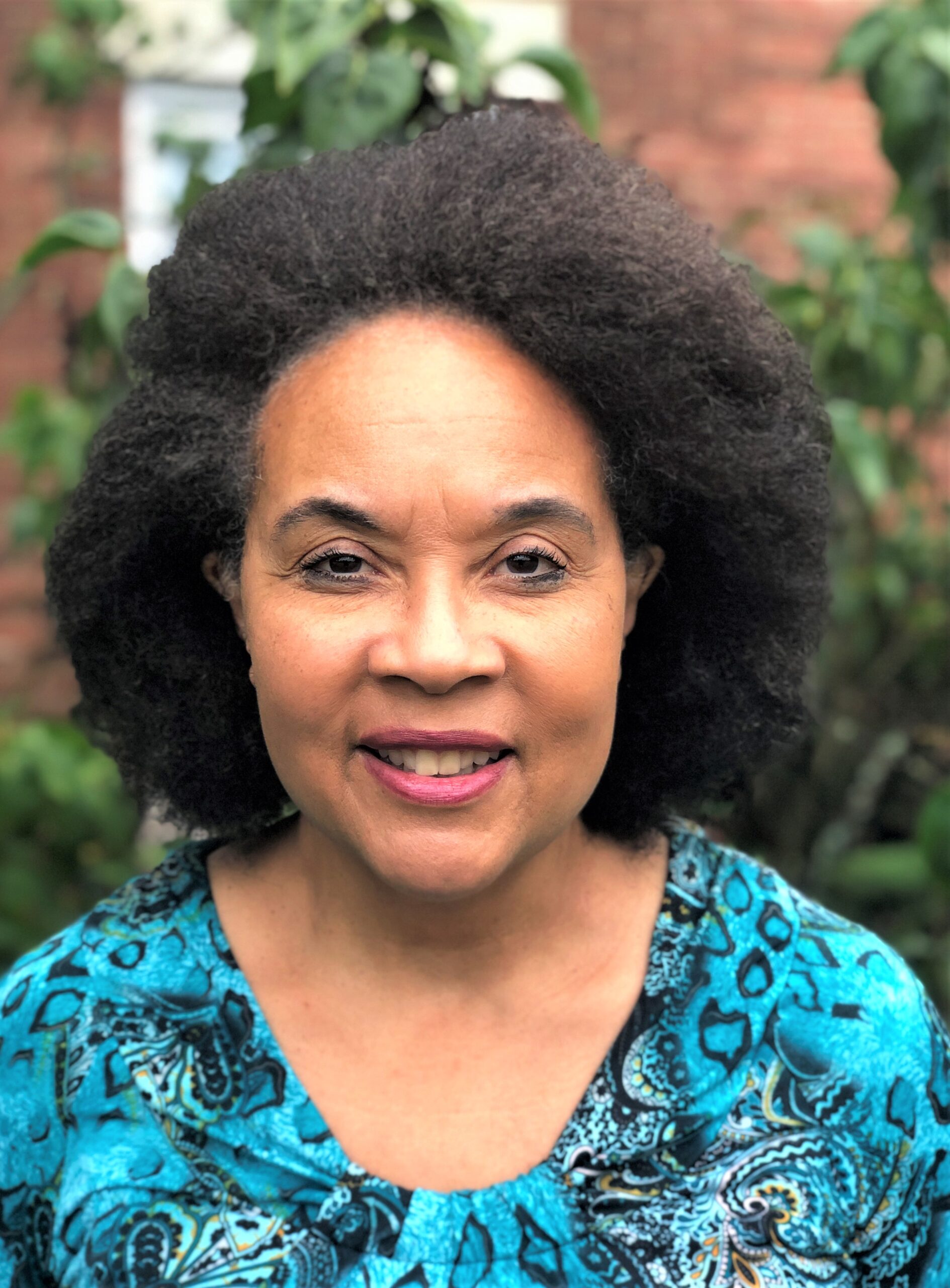 a woman standing outside in front of a brick building and green bush while wearing a brightly patterned blue shirt