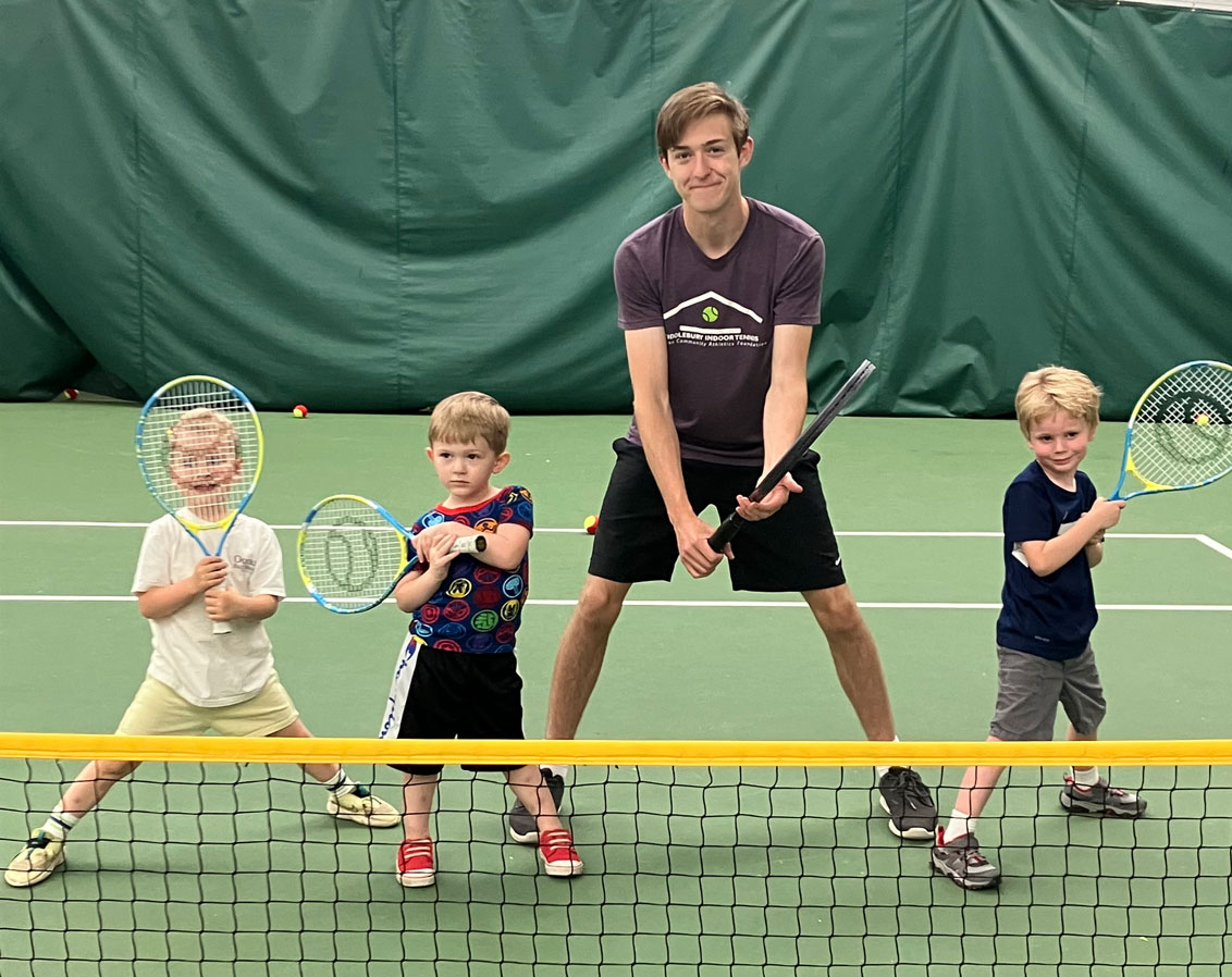 3 little kids and 1 teen holding tennis rackets and standing behind a tennis net