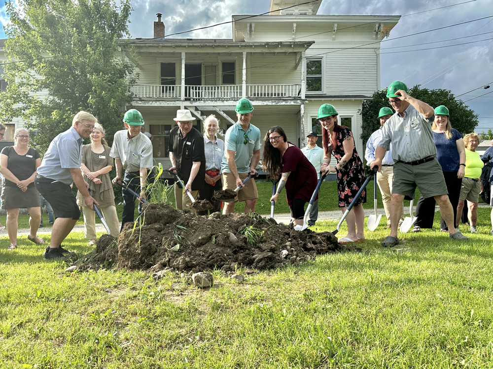 a group of mixed age people, many of them wearing green construction hats, in front of an older white building, digging into a pile of dirt at a groundbreaking ceremony for the start of a renovation project