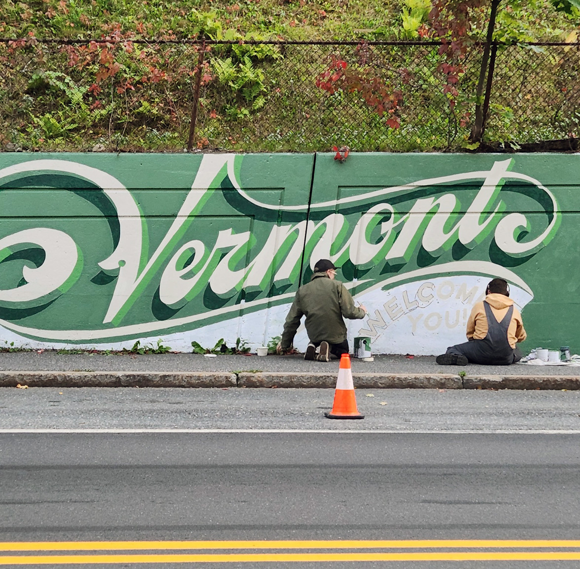 Two people work on a mural project on the side of a paved road.