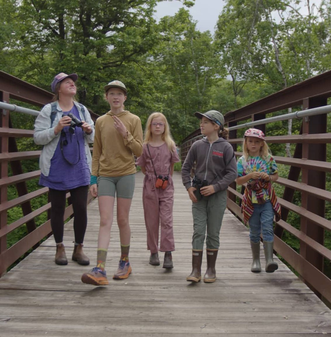 A group of five kids (of different ages) walk over a wooden bridge.
