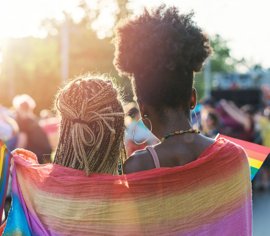 Two people look into the distance wrapped in a flag with rainbow colors.