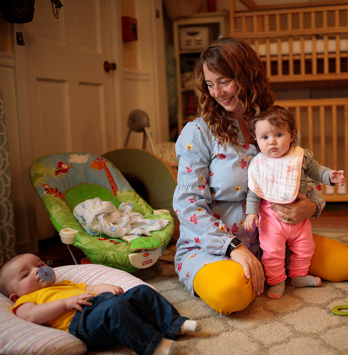 A woman sits on the floor in front of a row of cribs with two small children.