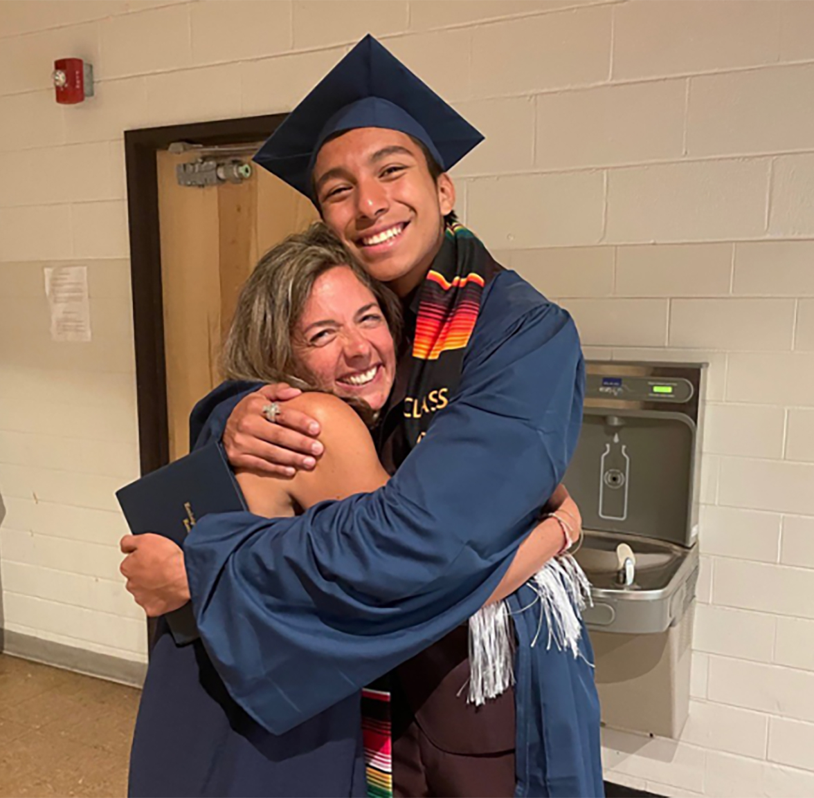 A teen in a graduation gown hugs his counselor.