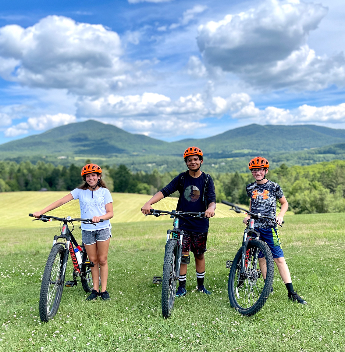 Three kids stand next to their bicycles with Vermont mountains in the distance.