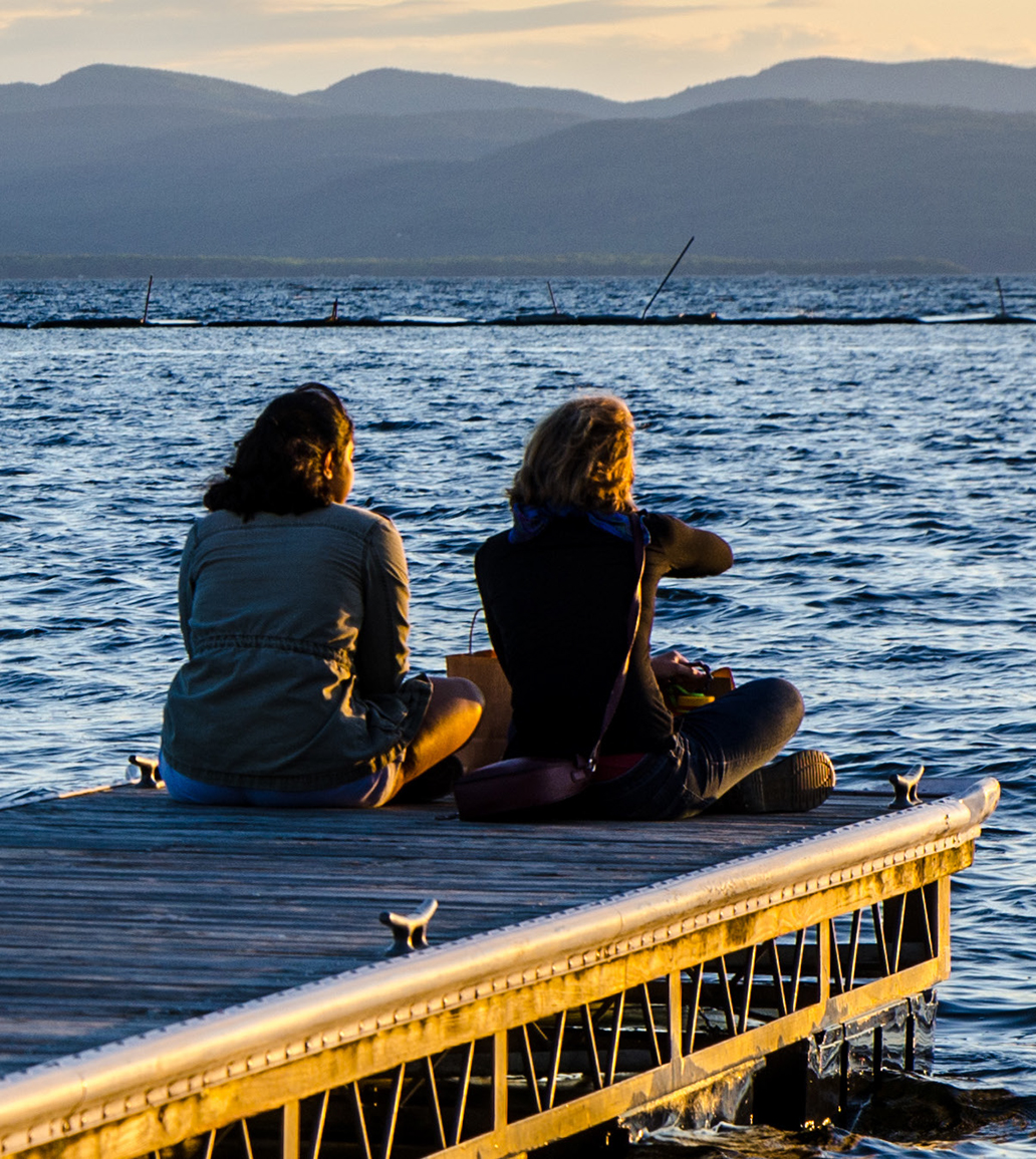 Two women sit on a dock on Lake Champlain chatting.