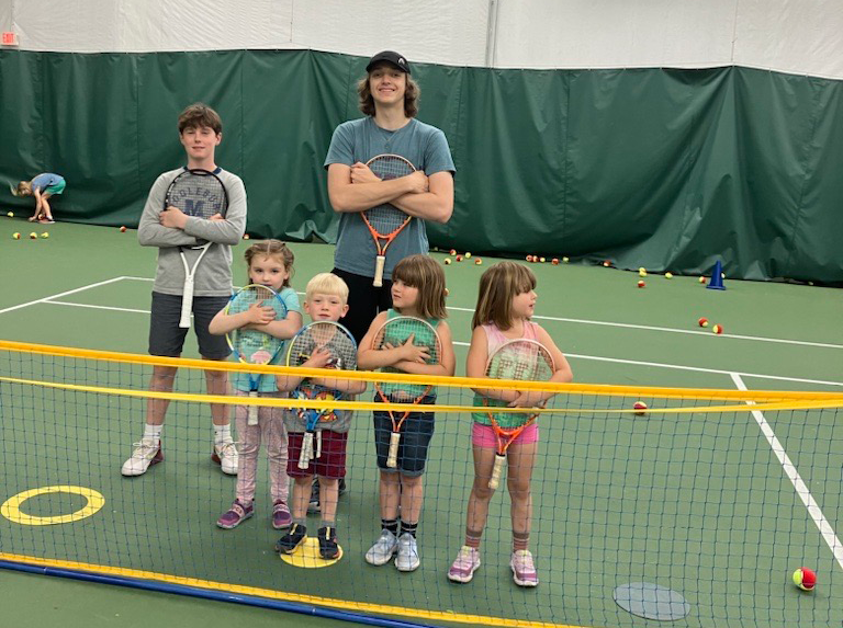 4 young kids holding tennis rackets, standing by a tennis net on a court, with 2 teen instructors standing behind them