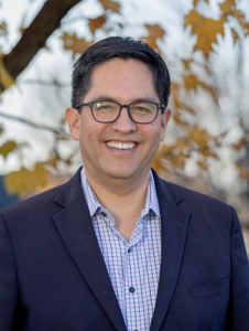 a man with dark hair in a blue button-up shirt and navy blazer