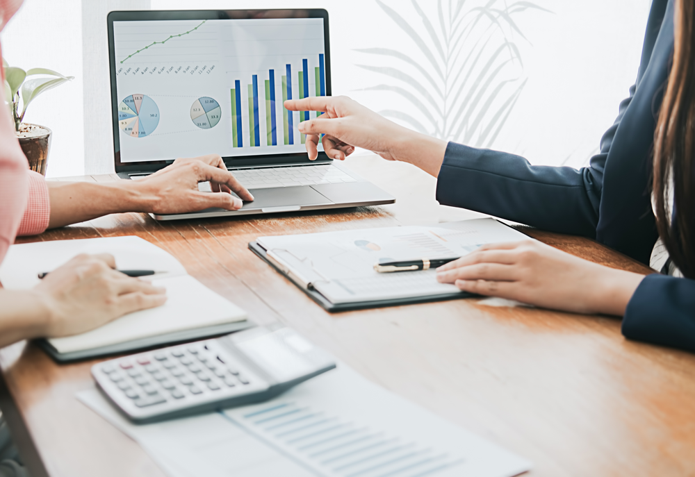 hands of 2 people across a desk with a calculator and a computer screen with charts & graphs