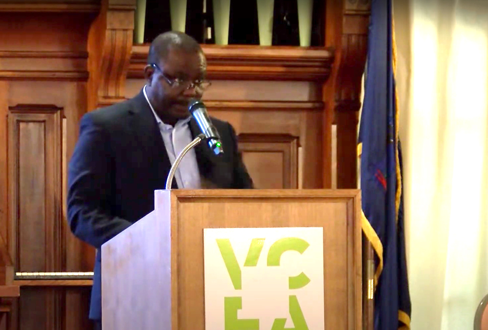 a man in a black suit with a white shirt speaking at a podium