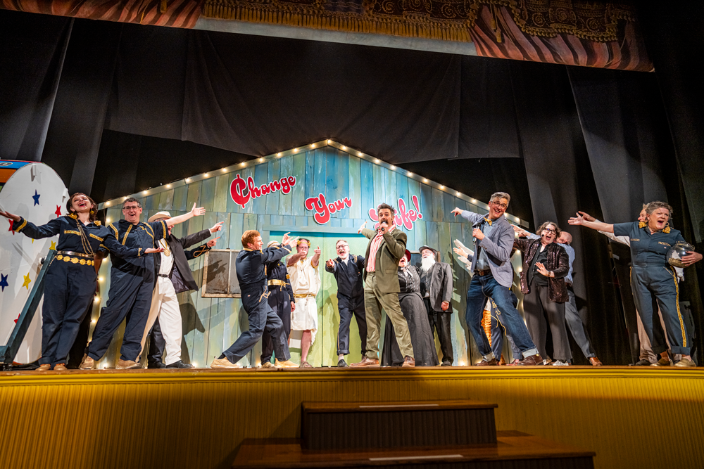 a group of smiley people on a stage pointing at the set background which reads 