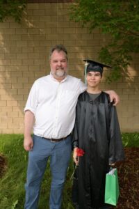 A graduate in their cap and gown proudly holds their degree with their father next to them.