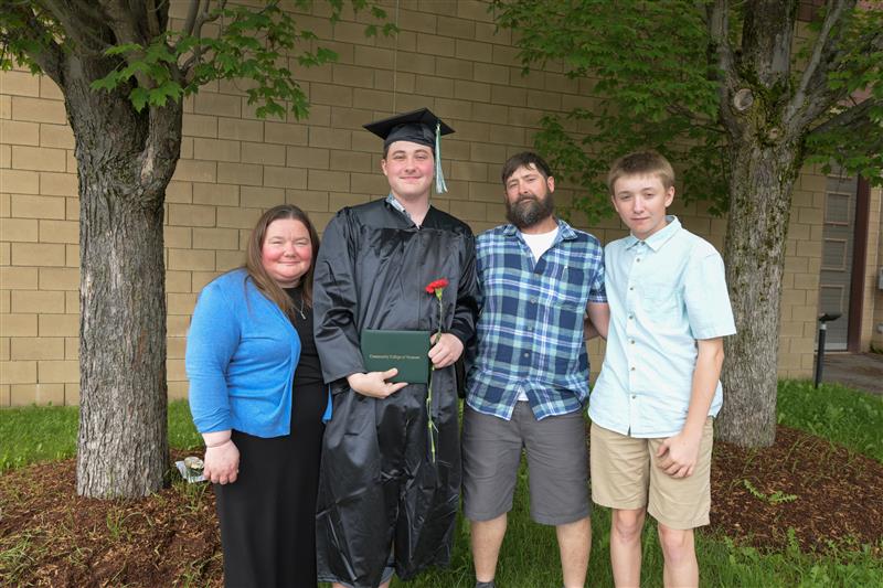 A graduate in their cap and gown proudly holds their degree with three family members around them.