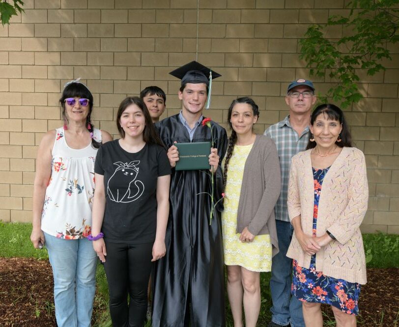 A graduate in their cap and gown proudly holds their degree with family members around them.
